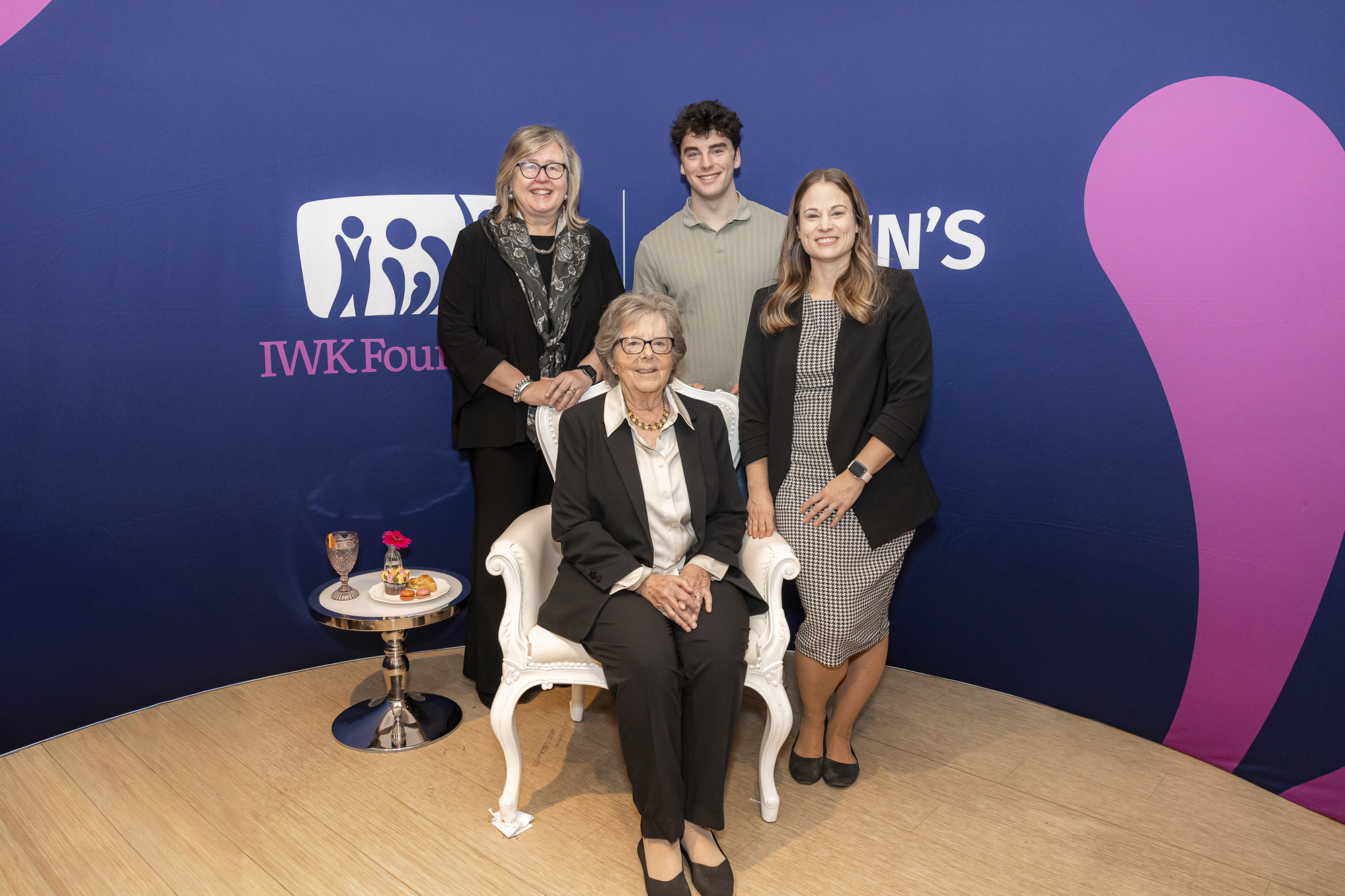 Group photo of four people at an IWK Foundation event with a seated woman in front and a small table holding a drink, snacks, and a flower.