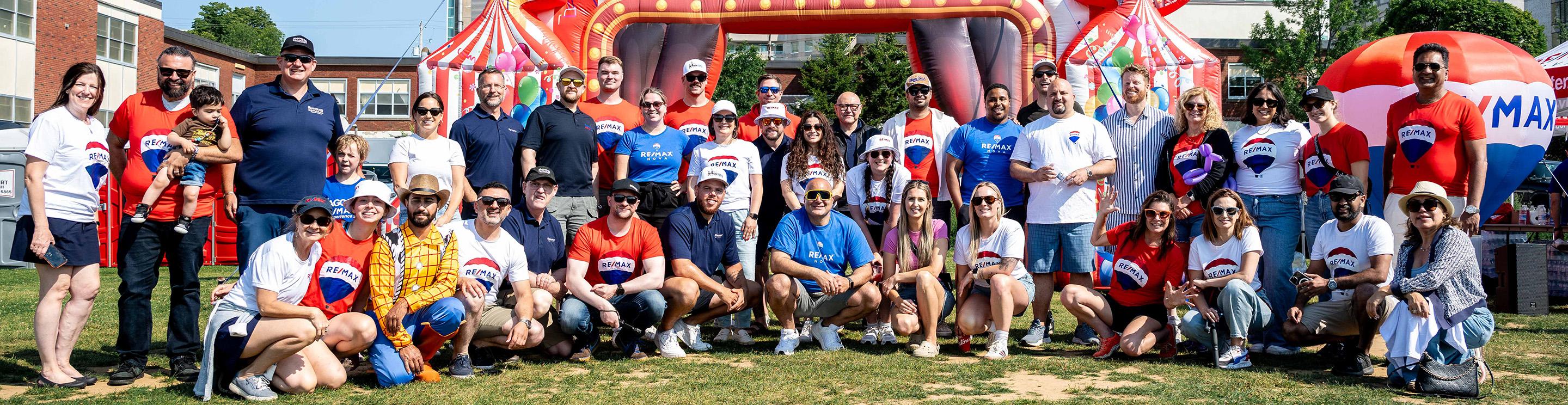 Group of RE/MAX agents posing together outdoors at a community event, wearing branded red, white, and blue shirts in front of colorful inflatable attractions.