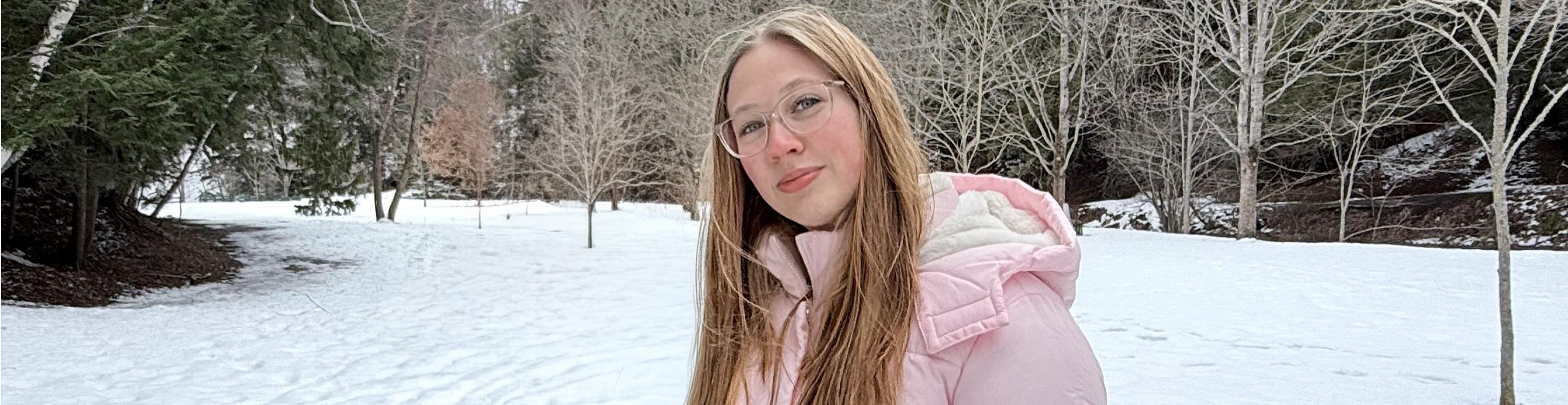 Brooke, wearing glasses and a light pink winter jacket, stands in a snowy park with bare trees and evergreen forest in the background.