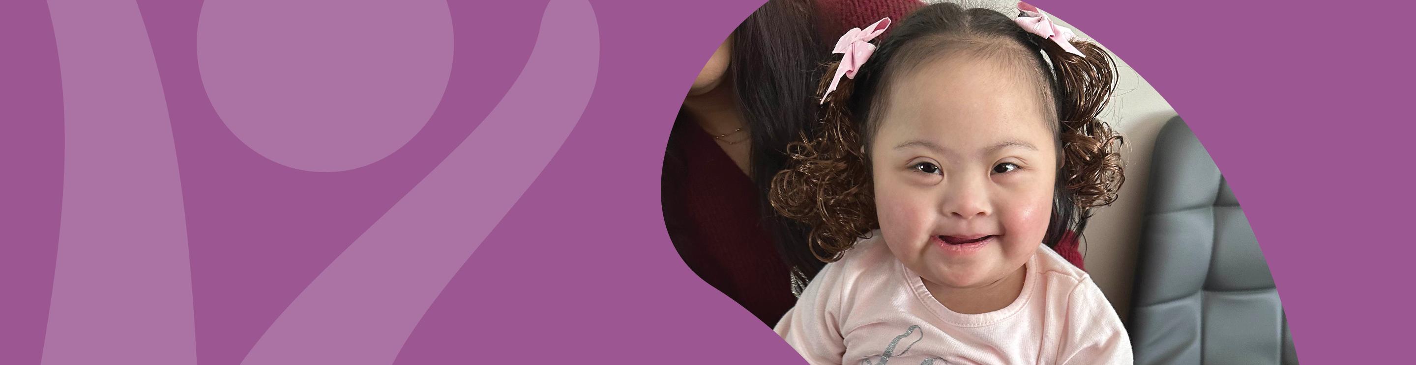 Juliana with pink bows in curly hair smiles while sitting with a family member.