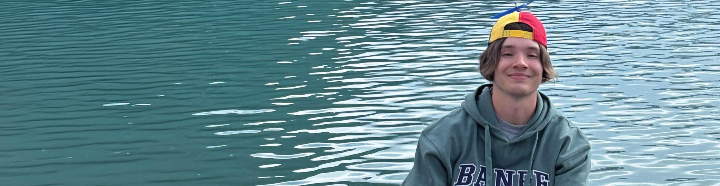 Quinn wearing a green hoodie with “BANFF” lettering and a multicolored backward cap sits near turquoise lake water. Gentle ripples reflect across the water’s surface, creating a serene outdoor setting.