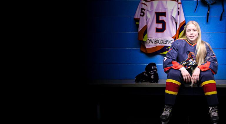 Young hockey player in navy, red, and yellow uniform sitting on a locker-room bench, skates on, with a number 5 jersey labeled “CDW BOOKKEEPING” hanging behind them; hockey gloves and stick nearby against a blue wall.