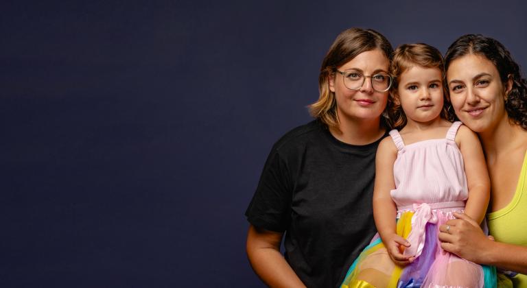 Studio portrait of two women and a toddler smiling together against a dark blue background, modern inclusive family with same-sex parents and young child.