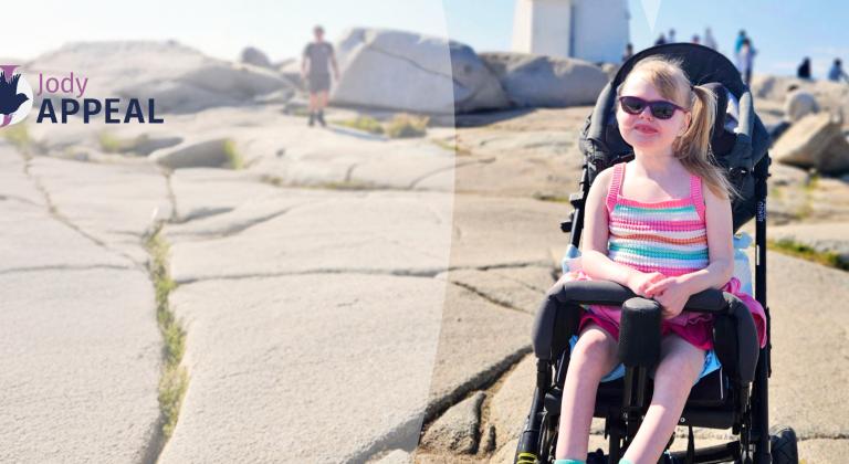 Lyla, an IWK patient, sitting in a stroller on a sunny rocky seaside path, with the “Jody Appeal” logo in the corner.