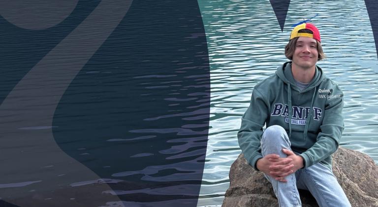 Quinn wearing a green hoodie with “BANFF” lettering and a multicolored backward cap sits near turquoise lake water. Gentle ripples reflect across the water’s surface, creating a serene outdoor setting.