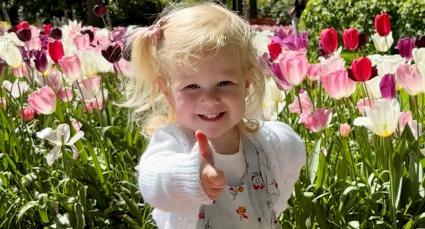 “Smiling young child giving a thumbs-up while standing among colorful pink, white, and purple tulips in a sunny garden.