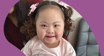 Juliana with pink bows in curly hair smiles while sitting with a family member.