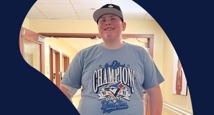 Landon wearing a Toronto Blue Jays championship T-shirt and baseball cap smiles while standing in a hallway at the IWK.
