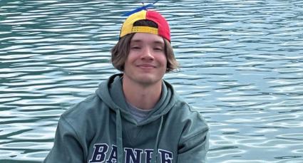 Quinn wearing a green hoodie with “BANFF” lettering and a multicolored backward cap sits near turquoise lake water. Gentle ripples reflect across the water’s surface, creating a serene outdoor setting.
