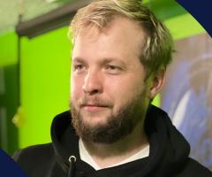 Alex Green wearing a black hoodie, photographed against a bright green and dark blue background with abstract graphic shapes.