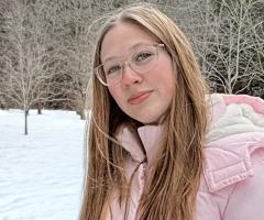 Brooke, wearing glasses and a light pink winter jacket, stands in a snowy park with bare trees and evergreen forest in the background.