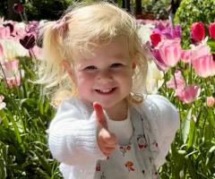 “Smiling young child giving a thumbs-up while standing among colorful pink, white, and purple tulips in a sunny garden.