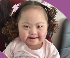 Juliana with pink bows in curly hair smiles while sitting with a family member.