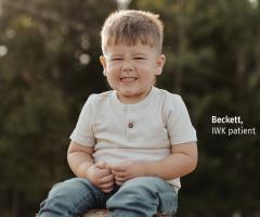 Beckett sitting outdoors in a wooded setting, smiling with his eyes closed and hands clasped in front of him. Text reads “Beckett, IWK patient.”