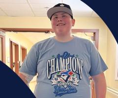 Landon wearing a Toronto Blue Jays championship T-shirt and baseball cap smiles while standing in a hallway at the IWK.