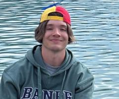 Quinn wearing a green hoodie with “BANFF” lettering and a multicolored backward cap sits near turquoise lake water. Gentle ripples reflect across the water’s surface, creating a serene outdoor setting.